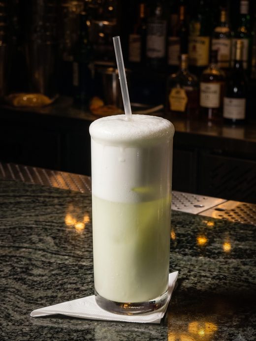 Um drink alto com espuma branca sobre o balcão de granito do bar, garrafas desfocadas atrás.