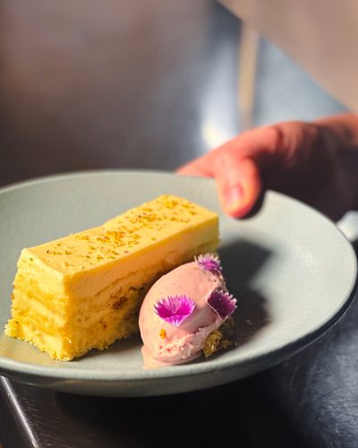 Fatia de bolo amarelo com uma quenelle de sorvete rosa e uma flor comestível.