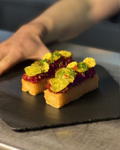 Duas fatias de pão tostado com creme roxo e flores amarelas, sobre uma ardósia.