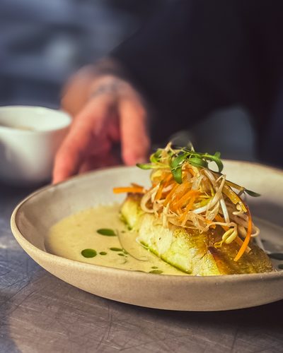 Filé de peixe com salada crocante por cima, num molho amarelo, prato fundo claro.