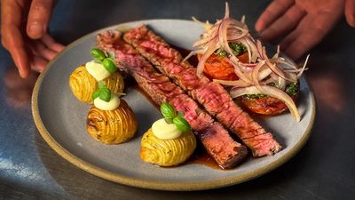 Vazio na brasa fatiado, com batata hasselback e cebola roxa, servido num prato azul.