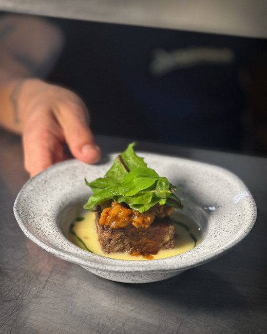 Um prato fundo claro sendo colocado na mesa, com carne braseada, purê e folhas verdes por cima.